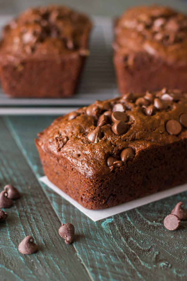 Chocolate Chocolate Chip Greek Yogurt Banana Bread loaf with chocolate chips sprinkled near it, and two more loaves on a cooling rack in the background.  