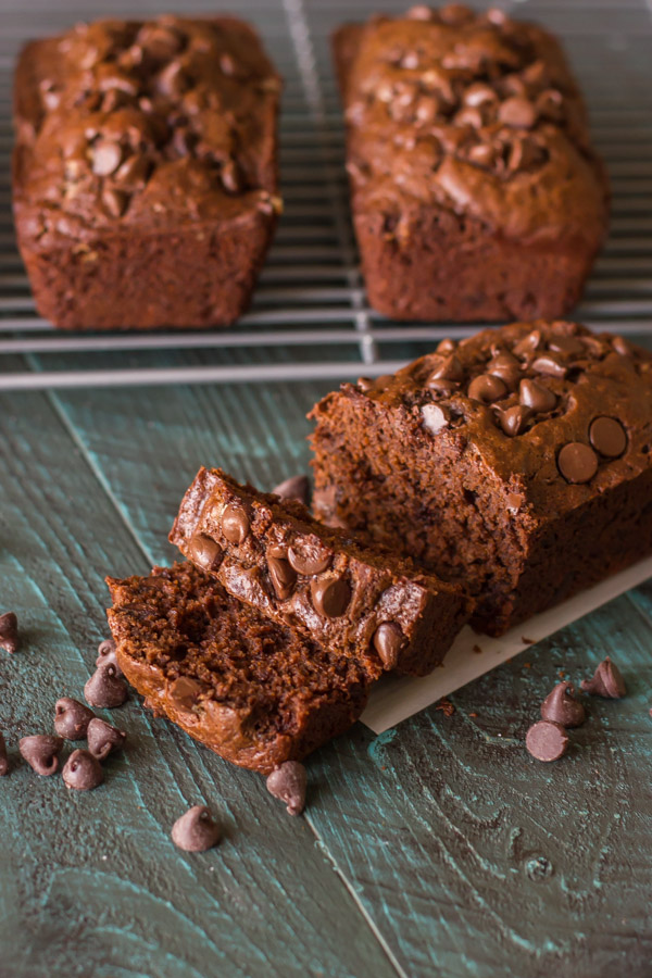 Chocolate Chocolate Chip Greek Yogurt Banana Bread loaf that has been sliced with chocolate chips sprinkled near it, and two more loaves on a cooling rack in the background.  