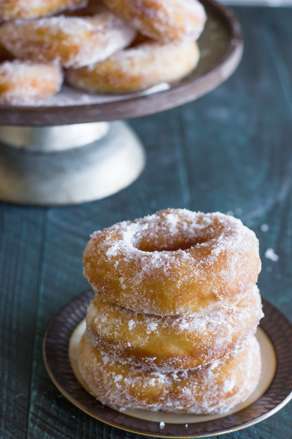 Homemade Yeast Doughnuts stacked in a pile of three on a plate, with more donuts on a cake stand in the background.