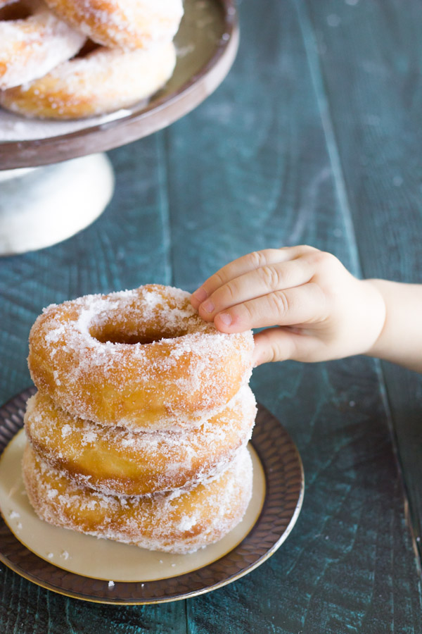 Homemade Yeast Doughnuts stacked in a pile of three on a plate with a hand grabbing the top donut and more donuts on a cake stand in the background.