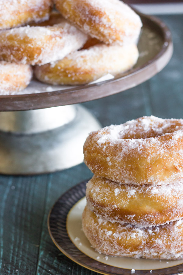 Homemade Yeast Doughnuts stacked in a pile of three on a plate, with more donuts on a cake stand in the background.
