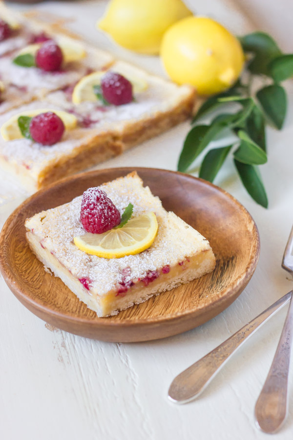 Raspberry Lemon Bar square serving on a wood plate, garnished with a lemon slice, a whole raspberry and a mint leaf. 
