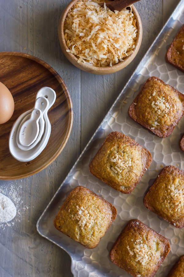 Whole Wheat Toasted Coconut Banana Muffins on a serving platter, with a bowl of toasted coconut sitting next to the platter, along with a wood plate with measuring spoons on it.  
