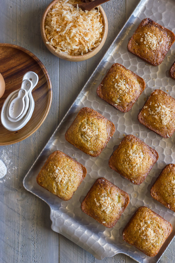 Whole Wheat Toasted Coconut Banana Muffins on a serving platter, with a bowl of toasted coconut sitting next to the platter, along with a wood plate with measuring spoons on it.  