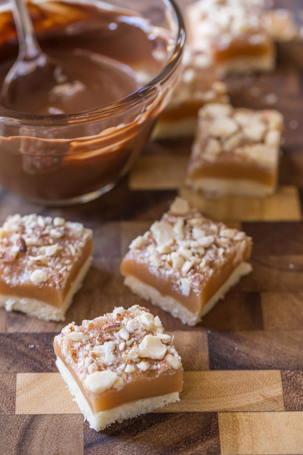 Homemade Caramel Almond Shortbread Bites on a cutting board with a glass dish of melted chocolate.