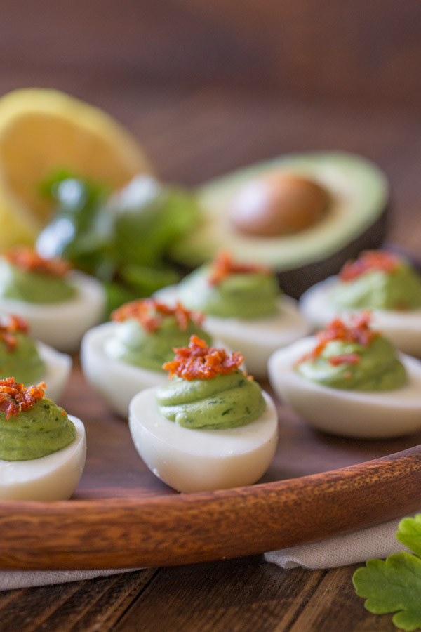 Smoky Bacon Avocado Deviled Eggs arranged on a wood serving plate, with a half of an avocado, a half of a lemon and a little bunch of cilantro.  