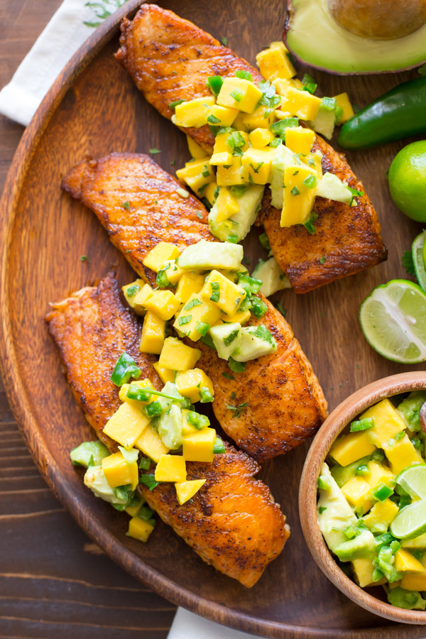 Chili Lime Salmon With Mango Avocado Salsa on a wood serving plate, with a half of an avocado, a jalapeño, a few limes, and a small wood bowl of the Mango Avocado Salsa.