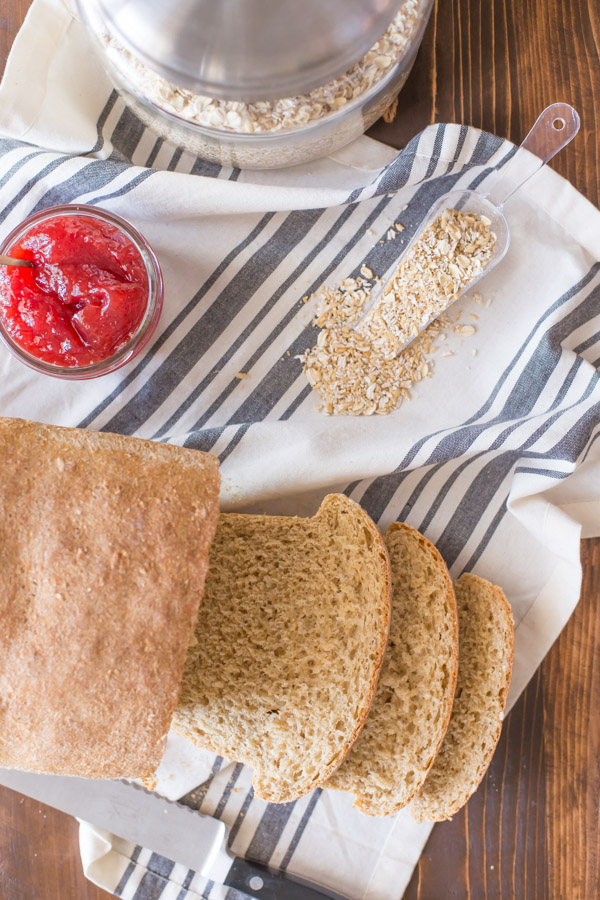 Easy Honey Whole Wheat Bread loaf that has been sliced, sitting on a cloth towel with oats and a jar of jelly.