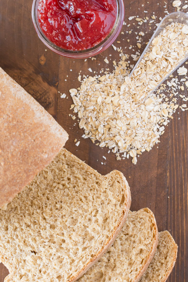 Easy Honey Whole Wheat Bread loaf that has been sliced, sitting next to some oats and a jar of jelly.