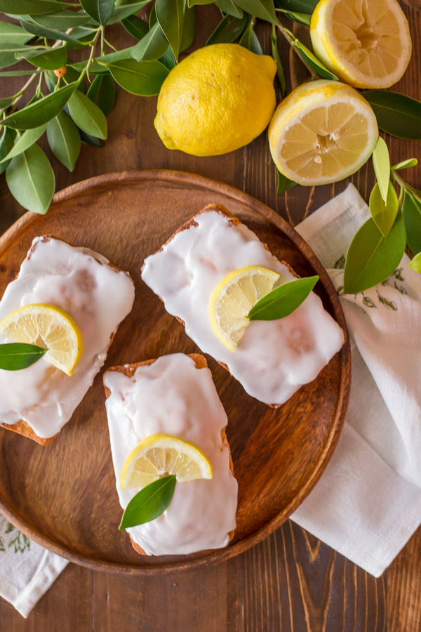 Mini Iced Lemon Pound Cake Loaves garnished with a lemon wedge and green leaf on a wood serving tray, next to some lemons and lemon tree leaves.