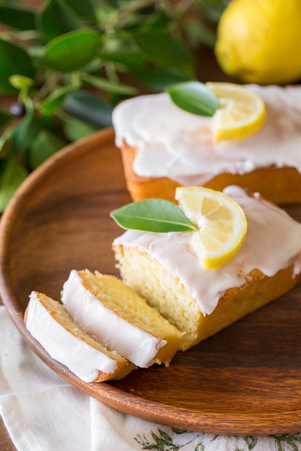 Mini Iced Lemon Pound Cake Loaf garnished with a lemon wedge and green leaf, that has two slices ready for serving, with another whole loaf sitting next to it on the same wood plate.