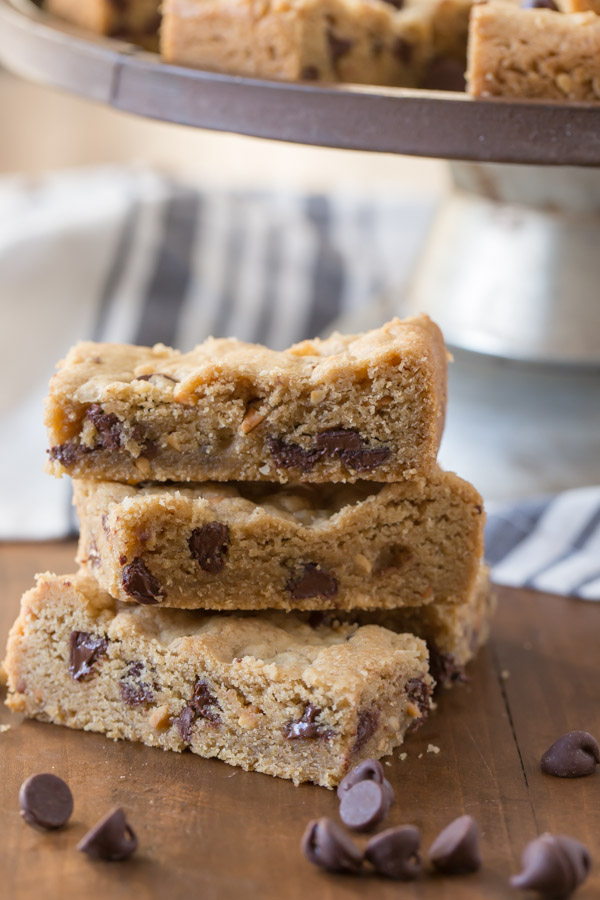 Peanut Butter Chocolate Chip Cookie Bars stacked in a pile, with more cookie bars on a cake stand in the background.  