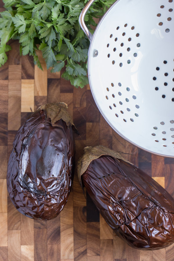 Two whole eggplants that have been broiled, sitting on a cutting board with a colander and some fresh parsley. 