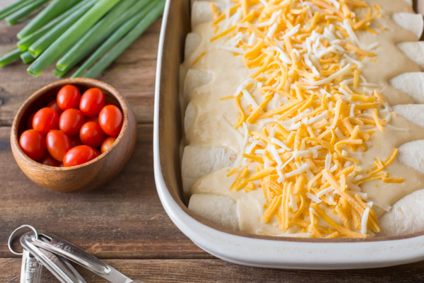 Breakfast Enchilada Bake assembled in a baking dish before being cooked, with grape tomatoes and green onions next to the baking dish. 
