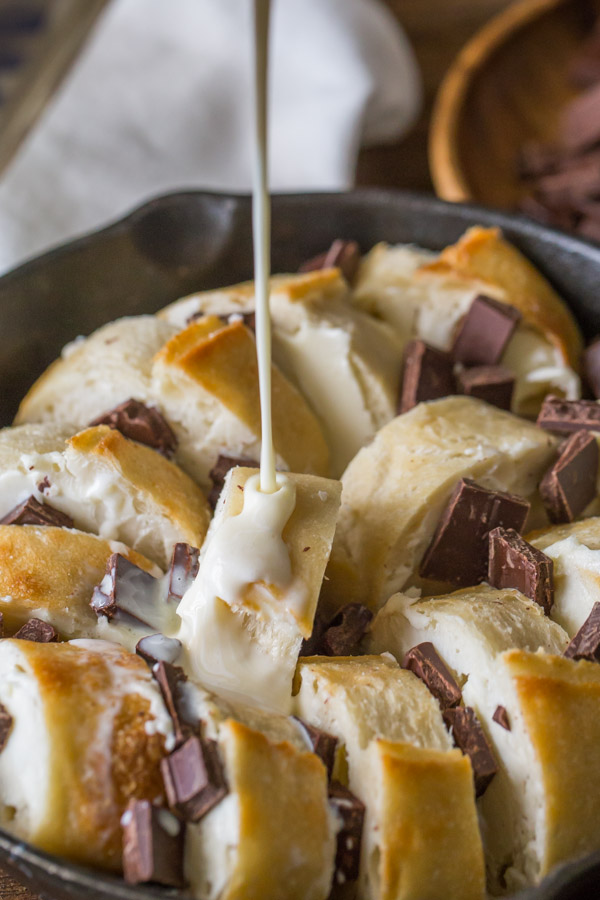 The creamy custard mixture being poured over the bread and chocolate chunks in the cast iron skillet. 