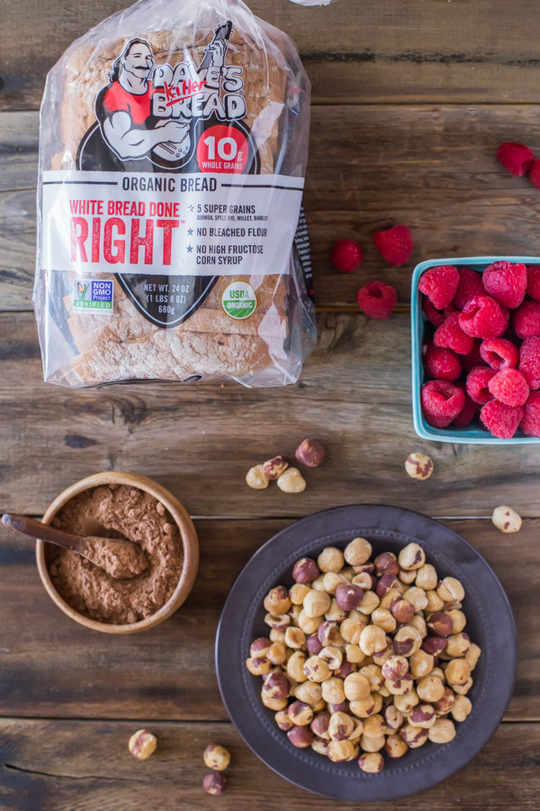 Some of the ingredients for Chocolate Hazelnut Raspberry Toast - a small bowl of cocoa powder, a plate of hazelnuts, a carton of raspberries and a bag of bread.