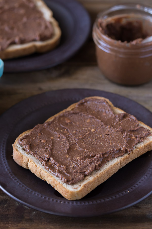 A piece of toast with Chocolate Hazelnut Butter spread on it, sitting on a plate, with a jar of Chocolate Hazelnut Butter in the background with another piece of toast with spread on it.