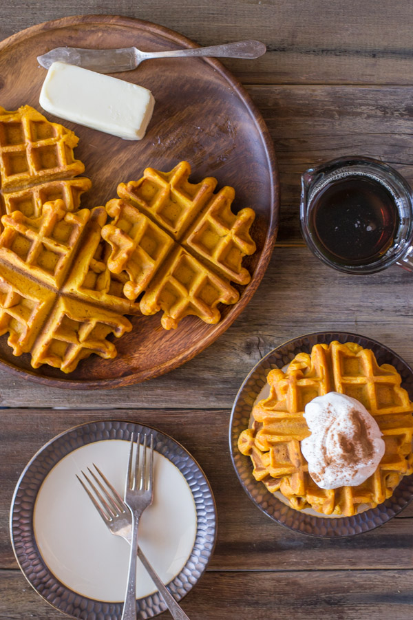 Pumpkin Spice Waffles on a wood serving plate with butter and a butter knife, sitting next to a glass container of maple syrup, a plate with a stack of Pumpkin Spice Waffles topped with whipped cream and cinnamon, and a plate with two forks on it.