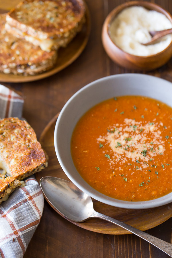 Homemade Tomato Soup in a bowl topped with grated Parmesan cheese, with a Garlic Bread Toasted Cheese Sandwich next to it, and more sandwiches on a wood plate in the background along with a small wood bowl of grated Parmesan cheese.