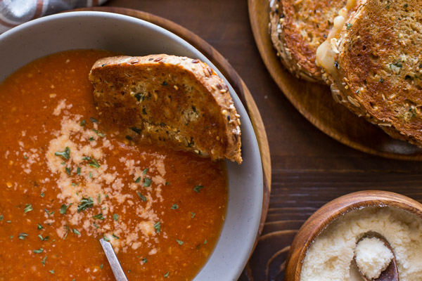 Homemade Tomato Soup in a bowl topped with grated Parmesan cheese with half a Garlic Bread Toasted Cheese Sandwich dipped in it, and more sandwiches on a wood plate next to it along with a small wood bowl of grated Parmesan cheese.