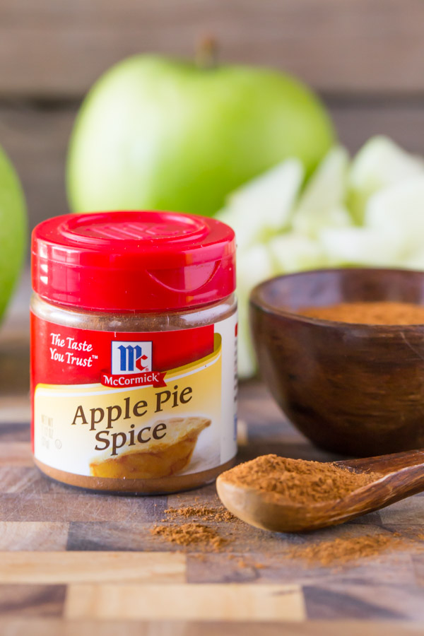 A container of McCormick Apple Pie Spice, sitting next to a small wood bowl of the spice and a small wooden spoon full of the spice. 