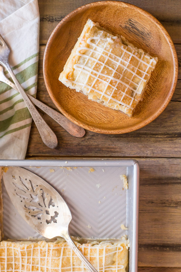 An Iced Apple Pie Bar on a wood plate, sitting next to the baking dish of Iced Apple Pie Bars with a serving spatula in it.    