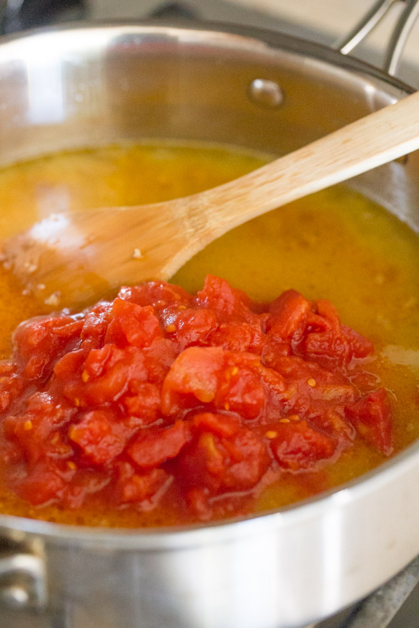 A large pan with a wooden spoon, showing the broth and tomatoes have been added for the One Pot Fusilli With Tomato, Basil, and Mozzarella.  