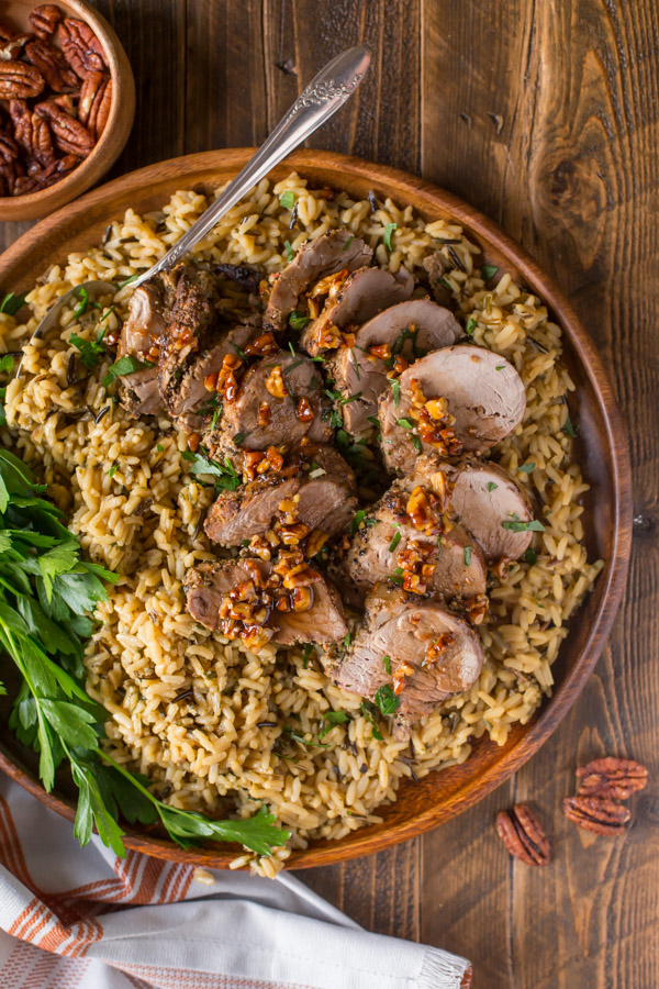 Pork Tenderloin sliced and served over rice and topped with a Sweet Chili Pecan Glaze on a wood plate with a serving spoon in it, with a small wood bowl of pecans next to it.