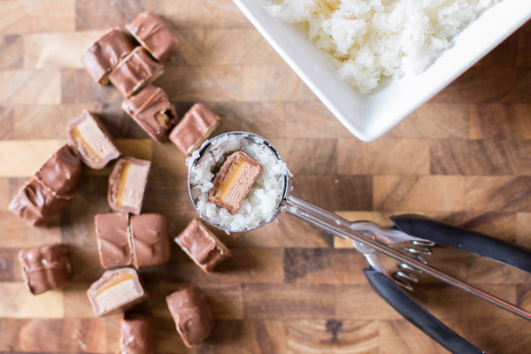 A cookie scoop with the coconut mixture packed into it along with a chocolate candy piece into the center, with a cutting board with candy bar pieces and a bowl of the coconut mixture in the background. 