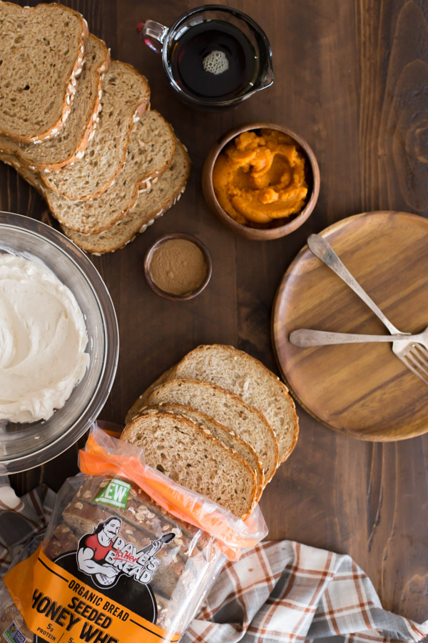 Pumpkin Pie Spiced Stuffed French Toast ingredients - a small wood bowl of pumpkin spice, a small wood bowl of pumpkin puree, a glass pour cup of maple syrup, slices of bread, a glass bowl of the cream cheese filling - sitting next to a plate with two forks.