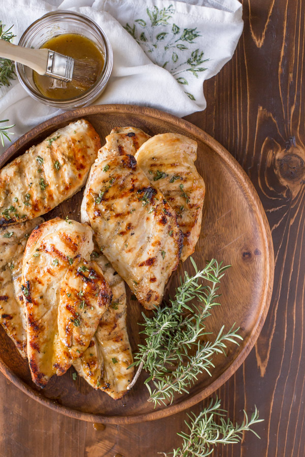 Honey Mustard Grilled Chicken breasts on a wood plate with some fresh rosemary, and a glass jar of the Honey Mustard Sauce with a basting brush in it sitting next to the plate. 