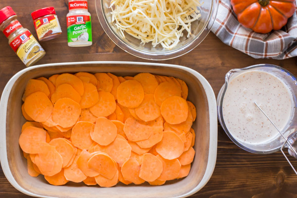 A baking dish with the sweet potato slices in it, sitting next to a glass cup of the heavy cream mixture, a bowl of shredded cheese and some spices.  