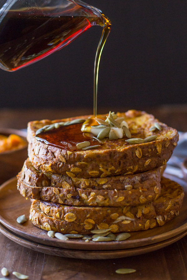 Pumpkin Pie Spiced Stuffed French Toast stacked on a wood plate, topped with butter and pepitas, with maple syrup being poured on top.
