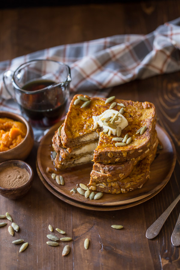 Pumpkin Pie Spiced Stuffed French Toast stacked on a wood plate and topped with butter, pepitas and maple syrup, with a section of the stack cut out. A small wood bowl of pumpkin spice, a small wood bowl of pumpkin puree, and a glass pour cup of maple syrup are sitting next to the plate.