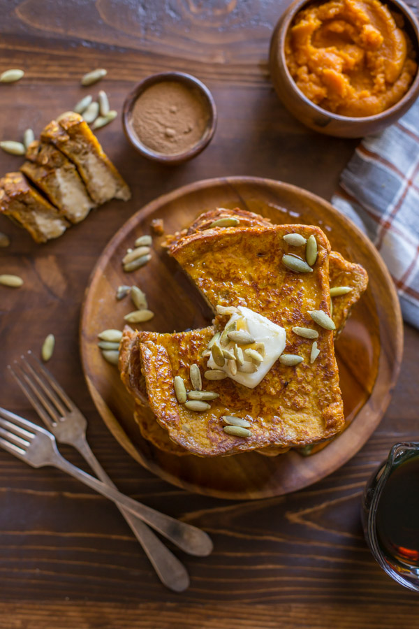 Pumpkin Pie Spiced Stuffed French Toast stacked on a wood plate and topped with butter, pepitas and maple syrup, with a section of the stack cut out and laying next to the plate, along with two forks, a small wood bowl of pumpkin spice, a small wood bowl of pumpkin puree, and a glass pour cup of maple syrup.