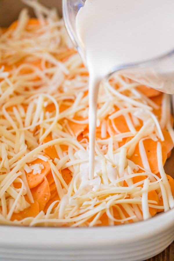 The heavy cream mixture being poured over the sweet potato slices that are topped with cheese for the Smoked Chili Sweet Potato Gratin.