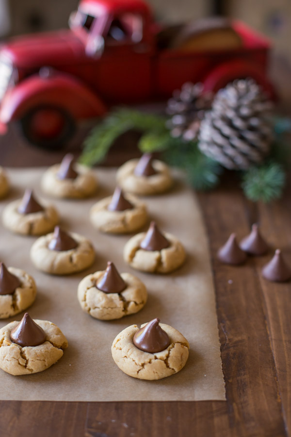 No Shortening Peanut Butter Blossoms sitting on a piece of parchment paper, with Hershey's Kisses next to them and Christmas decorations in the background.