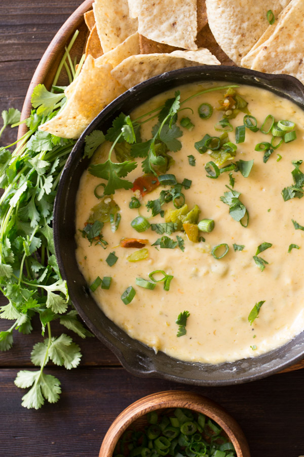 Hatch Green Chile Queso in a small cast iron skillet, garnished with green chiles, chopped cilantro, and green onions , sitting on a large wood plate with tortilla chips and a bundle of cilantro, with a small bowl of green onions next to the plate.  