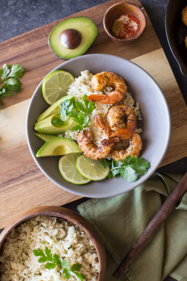 Cilantro Lime Rice Shrimp Bowl garnished with lime slices, avocado slices and fresh cilantro, sitting on a wood board with a small bowl of spices and a half of an avocado, and a wood bowl of Cilantro Lime Rice next to the board.