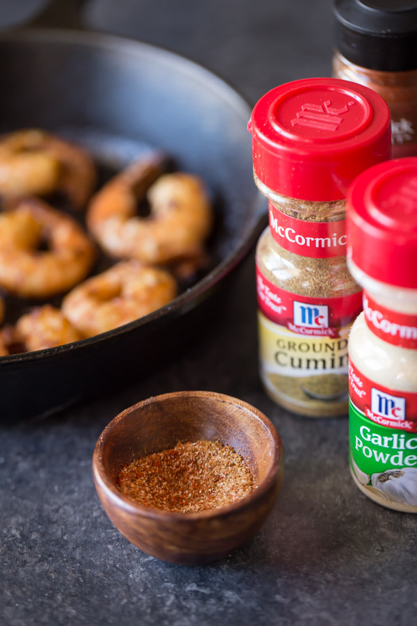 A small wood bowl of spices, with the spice containers and a cast iron skillet of shrimp in the background.