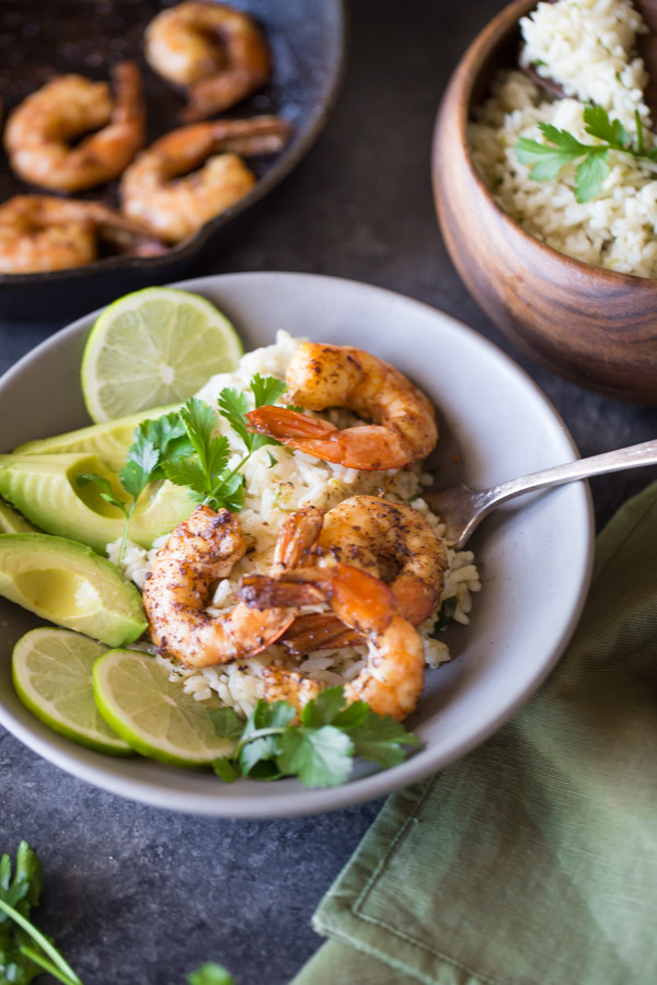 Cilantro Lime Rice Shrimp Bowl garnished with lime slices, avocado slices and fresh cilantro, with a wood bowl of Cilantro Lime Rice and a cast iron skillet of shrimp in the background.