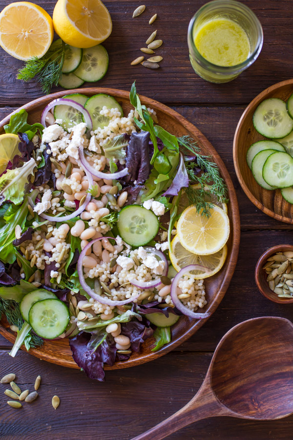 White Bean Couscous Salad With Lemon Vinaigrette on a large wood plate, with a wooden spoon, a small bowl of pepitas, a plate of sliced cucumbers, a glass jar of Lemon Vinaigrette, and a sliced lemon with some fresh dill and cucumber slices, all next to the plate of salad.