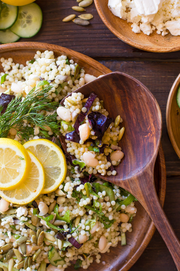 White Bean Couscous Salad With Lemon Vinaigrette on a large wood plate, garnished with lemon slices and fresh dill, with a wooden spoon in it and a wood bowl of feta cheese next to it.