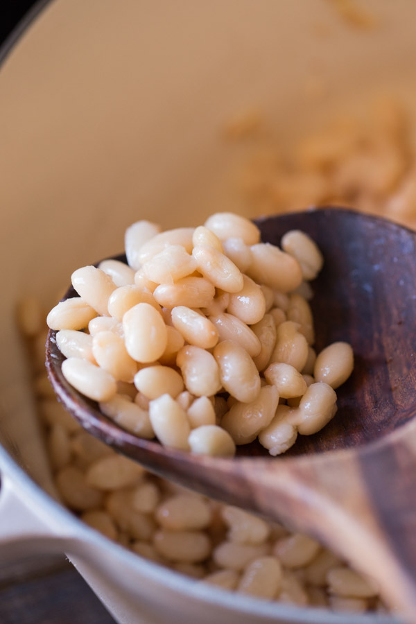 A wooden spoon full of white beans, being held over a large pot of white beans.