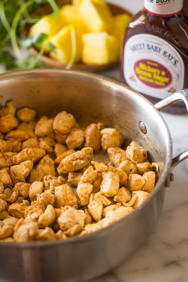 A large skillet with cooked chicken cubes in it, and a bottle of Sweet Baby Ray’s Hickory and Brown Sugar Barbecue Sauce and a plate of pineapple chunks in the background.