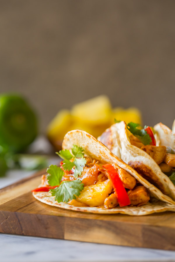 BBQ Chicken Fajitas in flour tortillas garnished with cilantro, on a cutting board, with a green pepper, fresh cilantro, and pineapple chunks in the background.