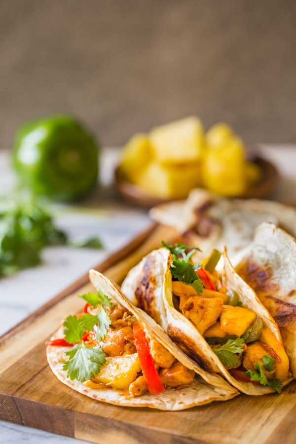 BBQ Chicken Fajitas in flour tortillas garnished with cilantro, on a cutting board, with a green pepper, fresh cilantro, and pineapple chunks in the background.