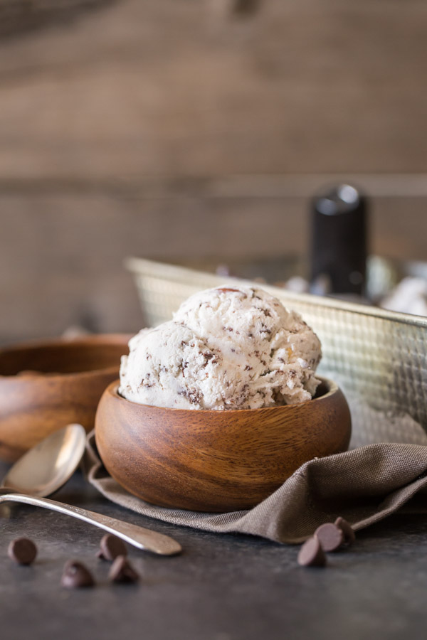 Dark Chocolate Almond Ice Cream in a small wood bowl, with a small wood bowl of almonds and a loaf pan of Dark Chocolate Almond Ice Cream in the background. 