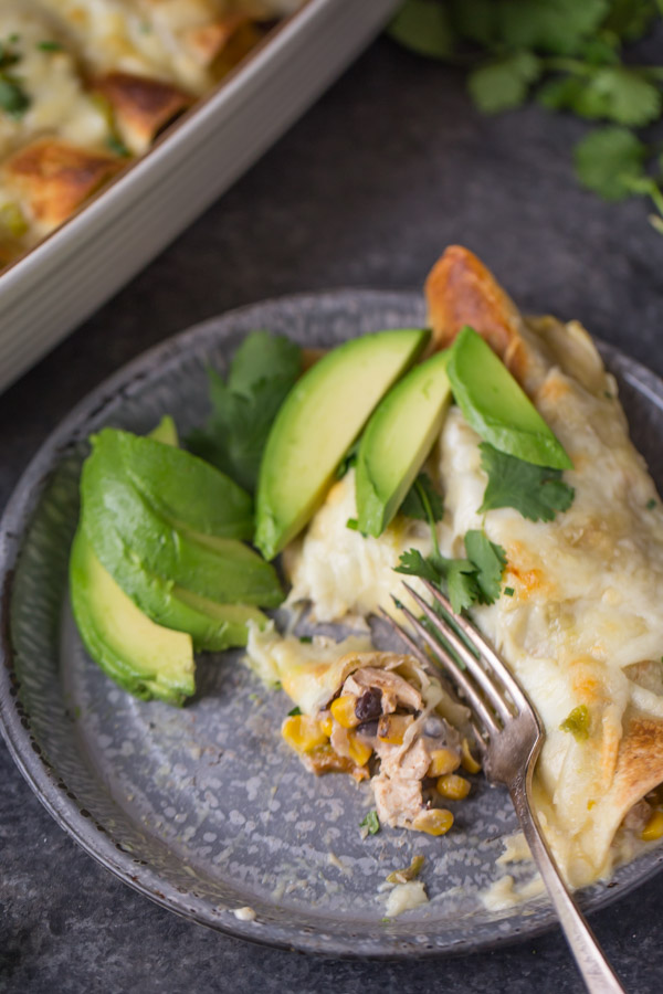 Hatch Green Chile Chicken Enchiladas on a plate with avocado slices, cilantro and a fork, sitting next to the baking dish of enchiladas.