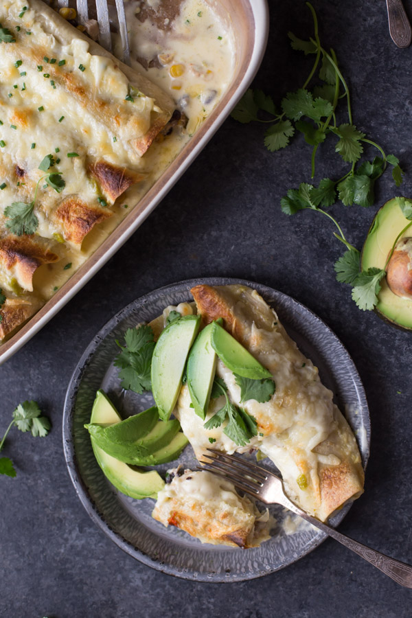 Hatch Green Chile Chicken Enchiladas on a plate with avocado slices, cilantro and a fork, sitting next to the baking dish of enchiladas.